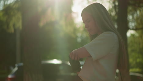 young adult wearing black gloves rubs elbow gently, sunlight filters through lush green leaves, casting a warm glow on her, blurred background with a passing car