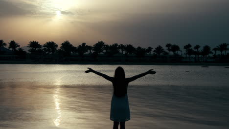 Beautiful-girl-silhouette-standing-arms-wide-open-at-sand-beach-in-sunrise.