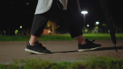 close-up leg view of person walking on night pathway holding umbrella, sneakers touching ground, casting shadow under streetlights, green grassy background