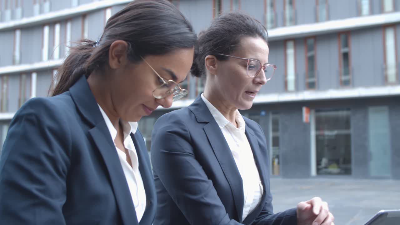 mujeres de negocios serias que usan computadoras portátiles mientras están sentadas cerca del edificio de oficinas afuera, hablando entre ellas