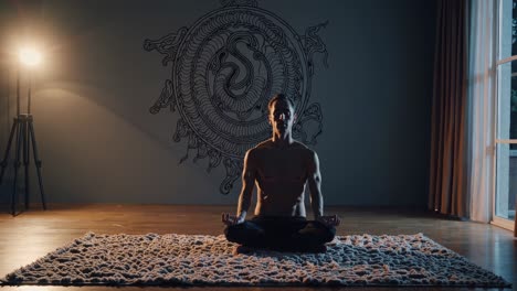 shirtless man sitting in lotus position on a rug in a dimly lit room, practicing yoga with a kundalini symbol drawn on the wall behind him, illuminated by a floor lamp