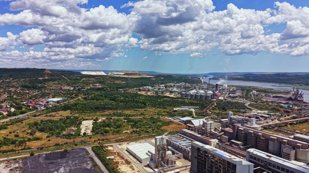 Zona industriale vicino al fiume con fabbriche, terreni agricoli e colline sotto un cielo nuvoloso durante il giorno.