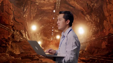 side view of an asian male professional worker standing in underground mine tunnel, observes by looking up before he come to concentrating on the laptop and keep on typing