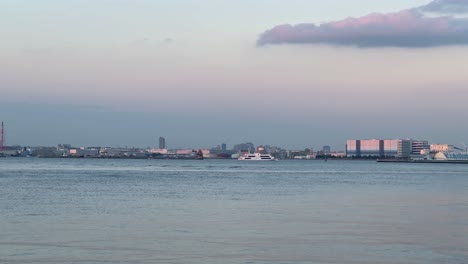 Pastel-sunset-over-a-calm-river-with-city-skyline-and-moving-boats,-soft-light