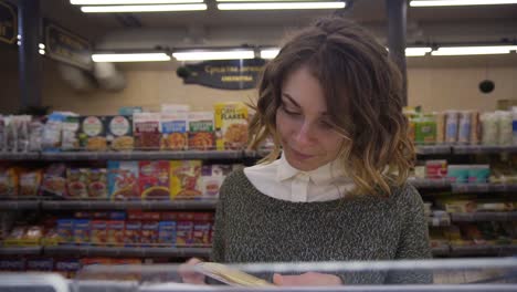 Young-curly-haired-woman-doing-grocery-shopping-at-the-supermarket,-she-is-reading-a-product-label-and-nutrition-facts-on-a-box-with-cheese.-Footage-from-the-shelf