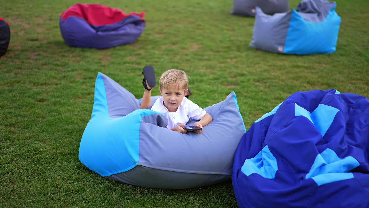 Green lawn with few bean bag chairs. Little blond boy lies on one of the chairs holding a smartphone in hands.