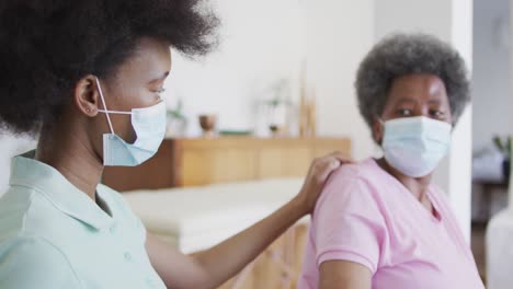 african american female physiotherapist wearing face mask helping senior female patient exercise