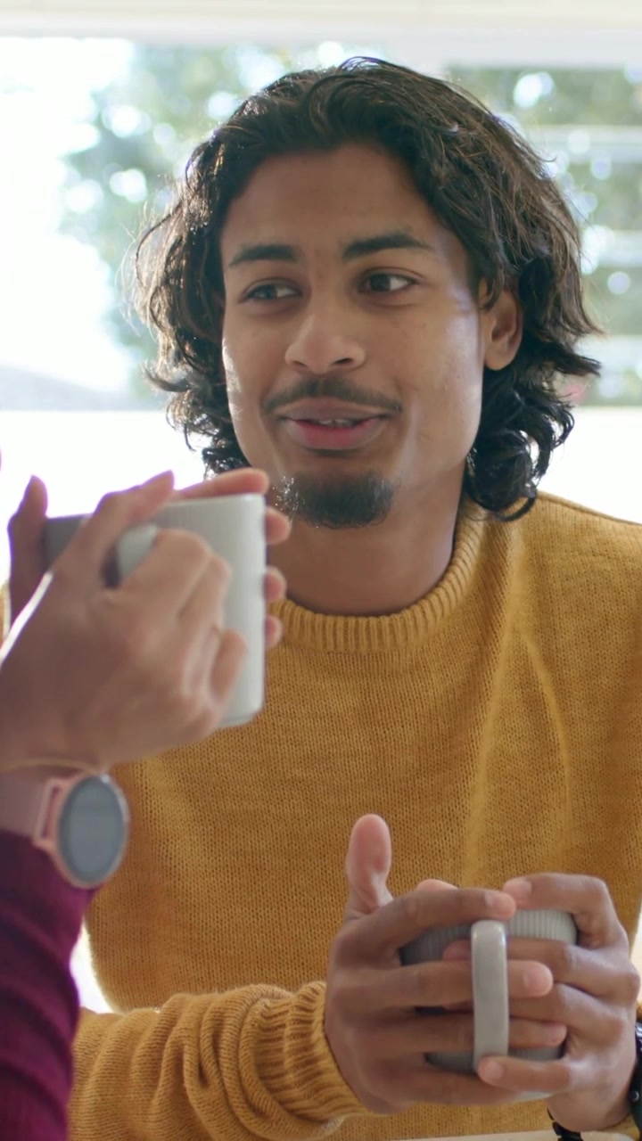 Video verticale: Uomo sorridente con maglia color senape che tiene una tazza e conversa in cucina mentre una donna alza una tazza.