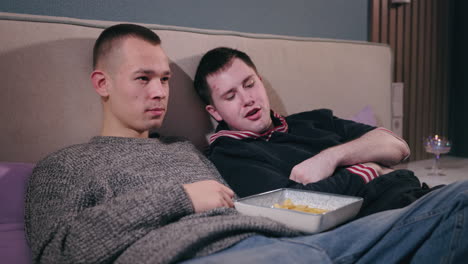 two men watching movie on couch and eating chips.