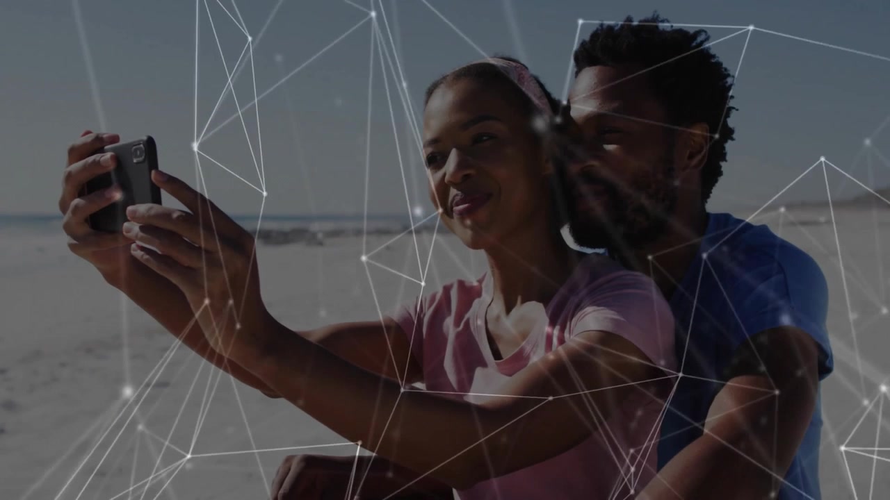Woman lifting smartphone and man embracing on beach generating network overlay for technology demo