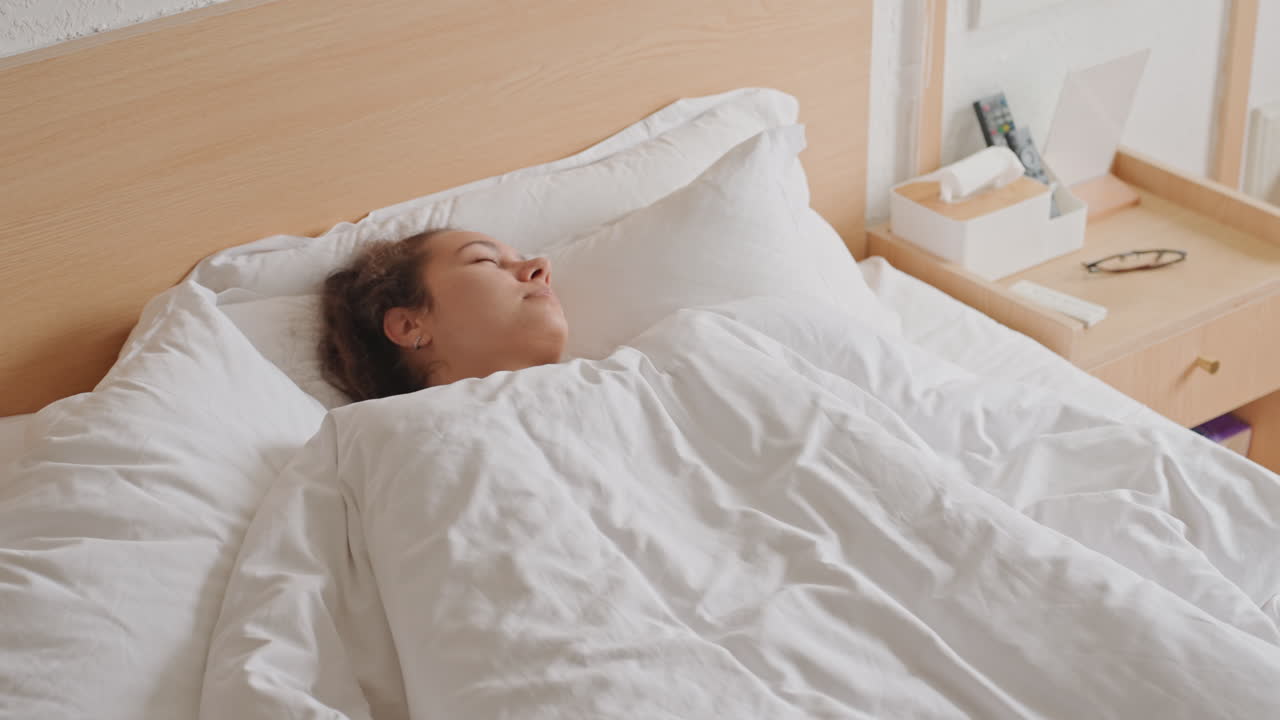 Ospite d'hotel irrequieta che combatte il jet lag, giovane donna sveglia di notte, controlla il telefono, sbadiglia e si sfrega gli occhi, si agita sotto il piumino, skyline della città visibile dalla finestra, valigia sul pavimento, tesa e stanca.