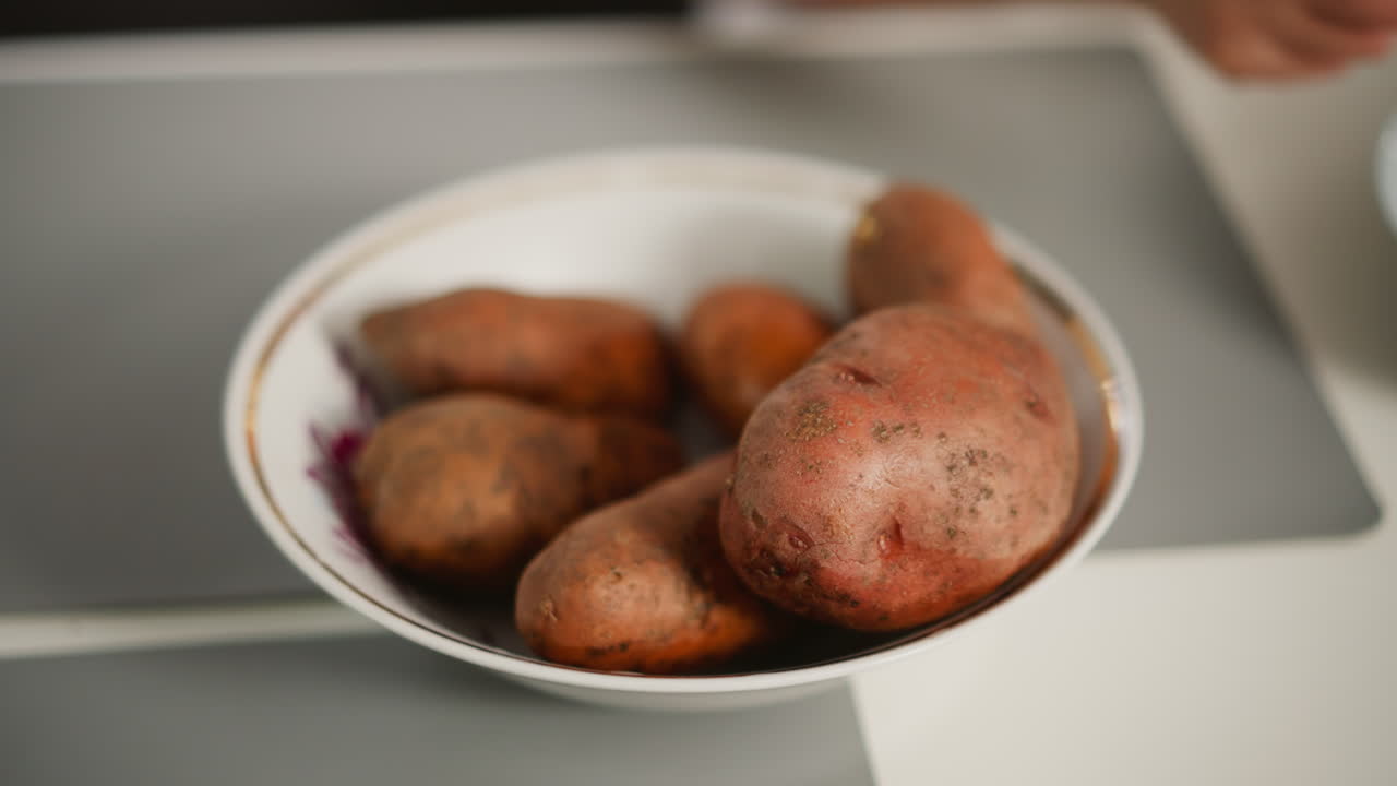 Close up of fair skinned person reaching into glass plate to retrieve raw potato, hand gently touching top of earthy vegetables in bowl placed on kitchen table with soft natural light and muted tones