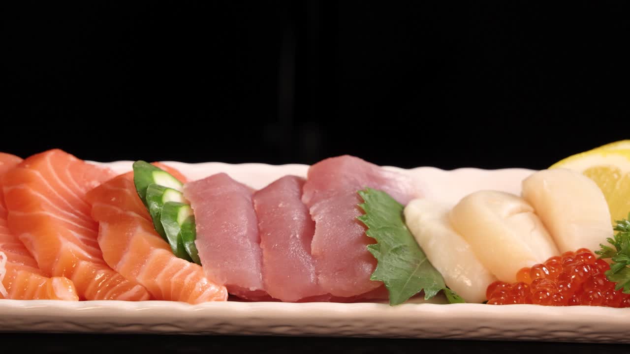 A rectangular plate of assorted sashimi, including salmon, tuna, scallop, and salmon roe, rotates smoothly against a black background under soft studio lighting