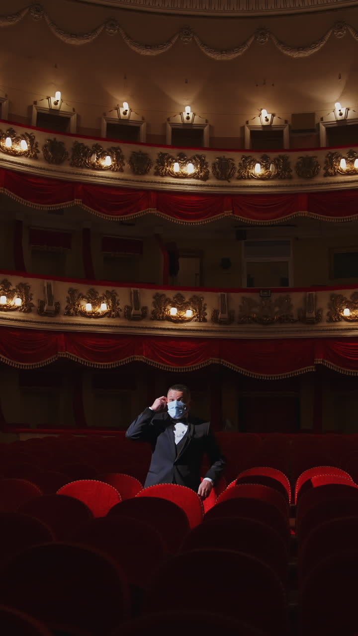 Spectator in mask sitting alone in empty theater. Man putting off his facial mask and talking in large dark auditorium among many rows of vacant red chairs in the theater. Vertical video