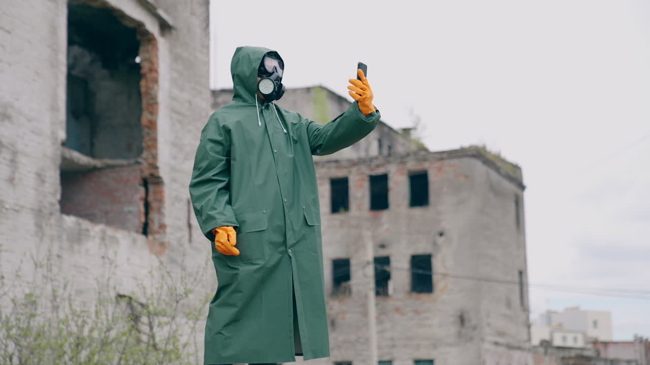 Human in a protective suit and gas mask with a phone. Unrecognizable person in safety uniform, gloves and respirator doing selfie in the destroyed place during radiation.