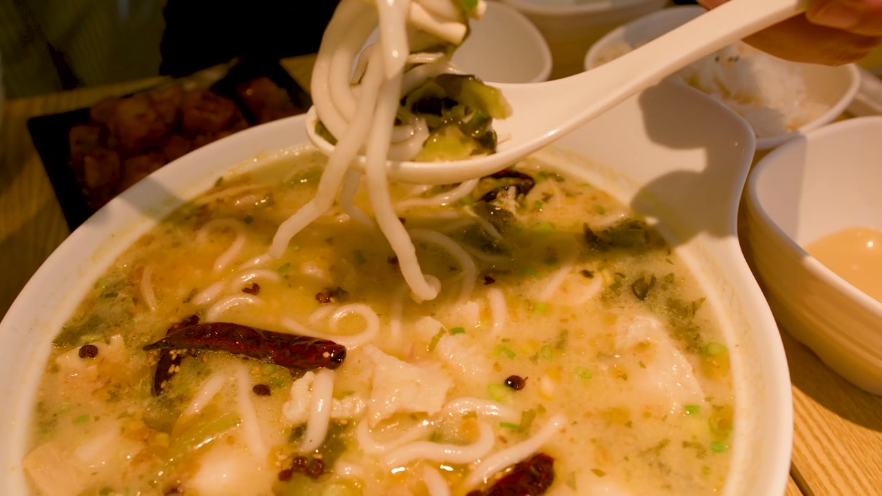 Primo piano di una persona che usa un cucchiaio e le bacchette per mangiare una zuppa piccante di noodle di riso.