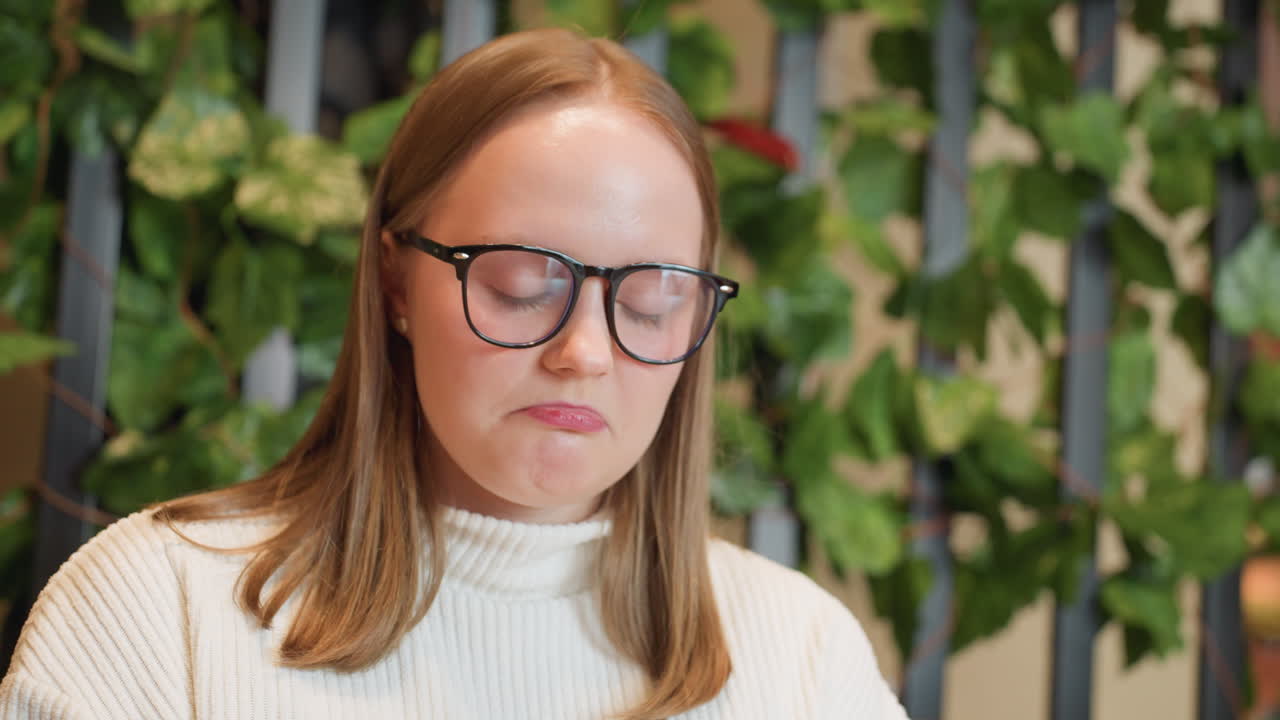 woman in glasses holds plate of chocolate cake near face, smelling dessert with contemplative expression in wooden cafe setting with green plants behind