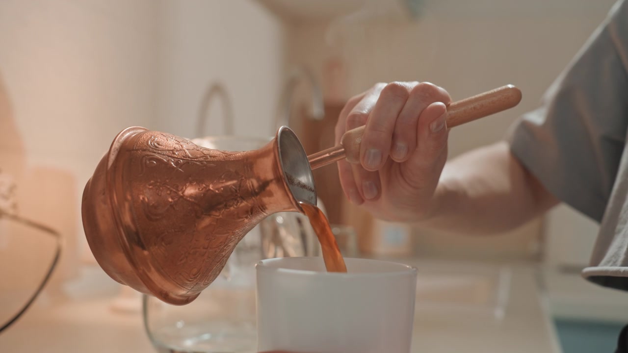 Closeup pouring Turkish coffee into mug, copper cezve and wooden stirrer on kitchen countertop with soft lamp glow intimate morning ritual, careful stirring and foam forming, home musician savoring