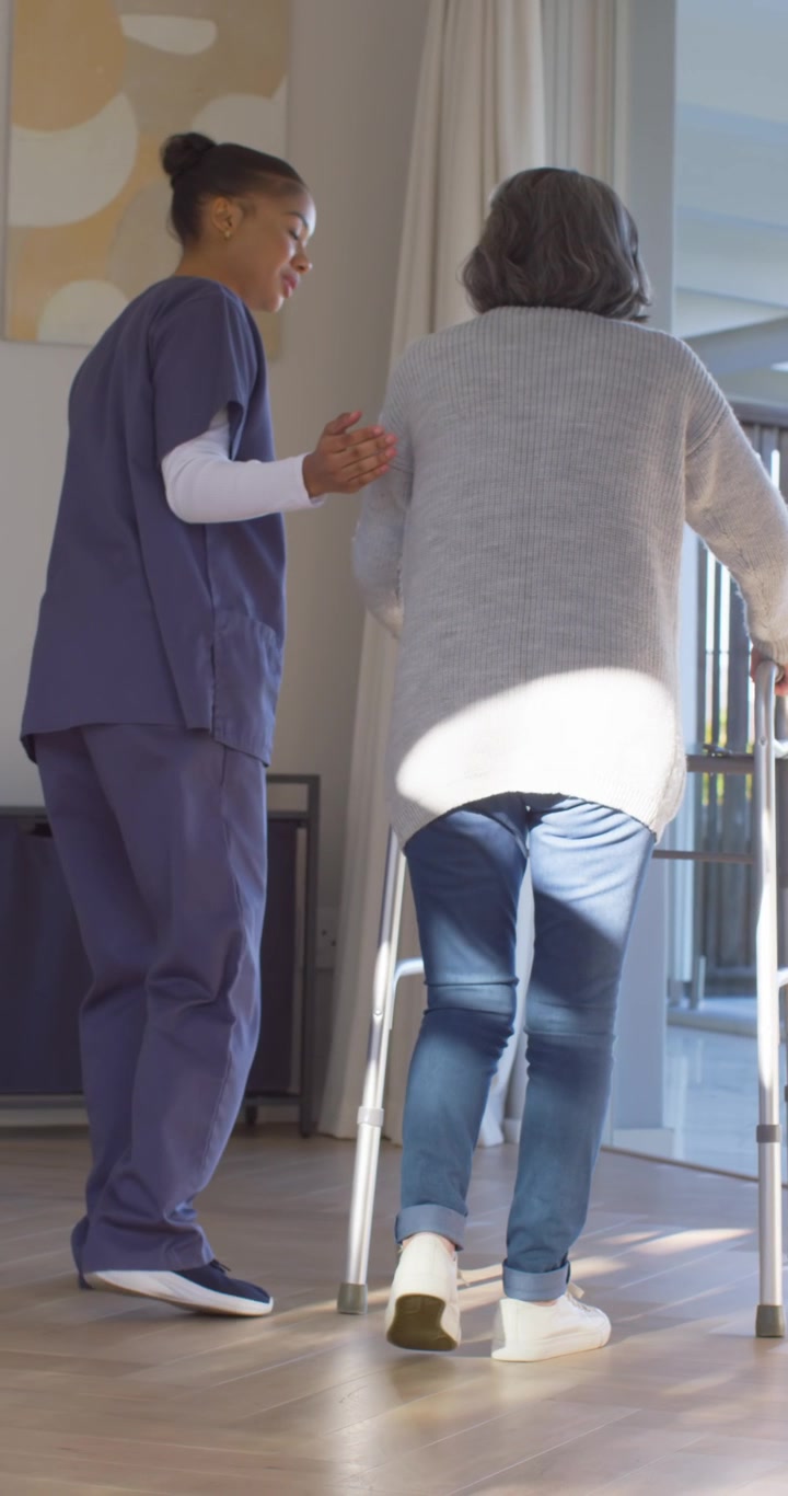 Vertikal video: Sykepleier i uniform veileder eldre person med rullator, hånd på ryggen, øver på gange hjemme