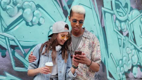 hipster couple in stylish clothes watching something on smartphone near a grafitti wall in the street