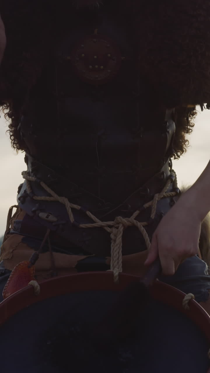 Drummer in leather vest with fur taps tambourine against distant mountains at sunset closeup. Man performs Altai music at ethnic festival