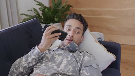 Glücklicher-Biracial-Männlicher-Soldat-In-Uniform,-Der-Zu-Hause-Auf-Dem-Sofa-Liegt-Und-Auf-Dem-Smartphone-Spricht,-Zeitlupe