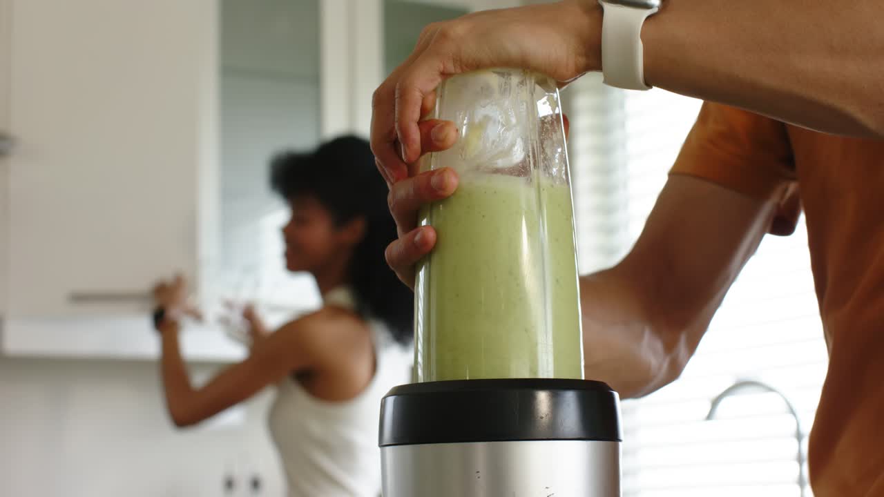 Pasangan beragam, pria meletakkan blender di atas dasar sambil memegang smoothie yang sudah diblender untuk disajikan kepada wanita yang memegang gelas