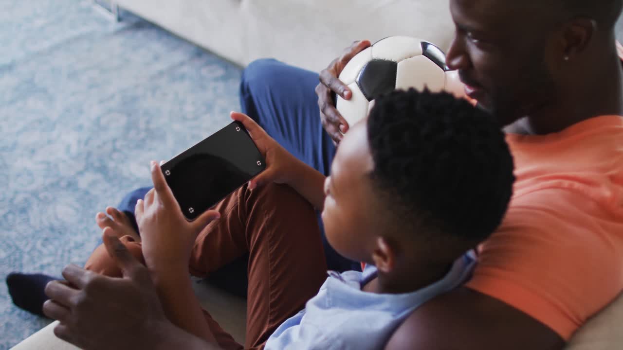 African american father and son using smartphone with copy space while sitting on the couch at home