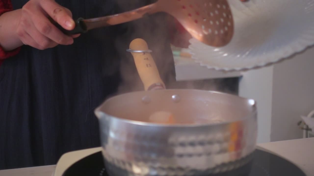 Close-up of shrimp being transferred from a plate into a steaming pot using a copper slotted spoon in a warm indoor kitchen.