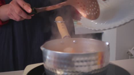 close-up of shrimp being transferred from a plate into a steaming pot using a copper slotted spoon in a warm indoor kitchen.