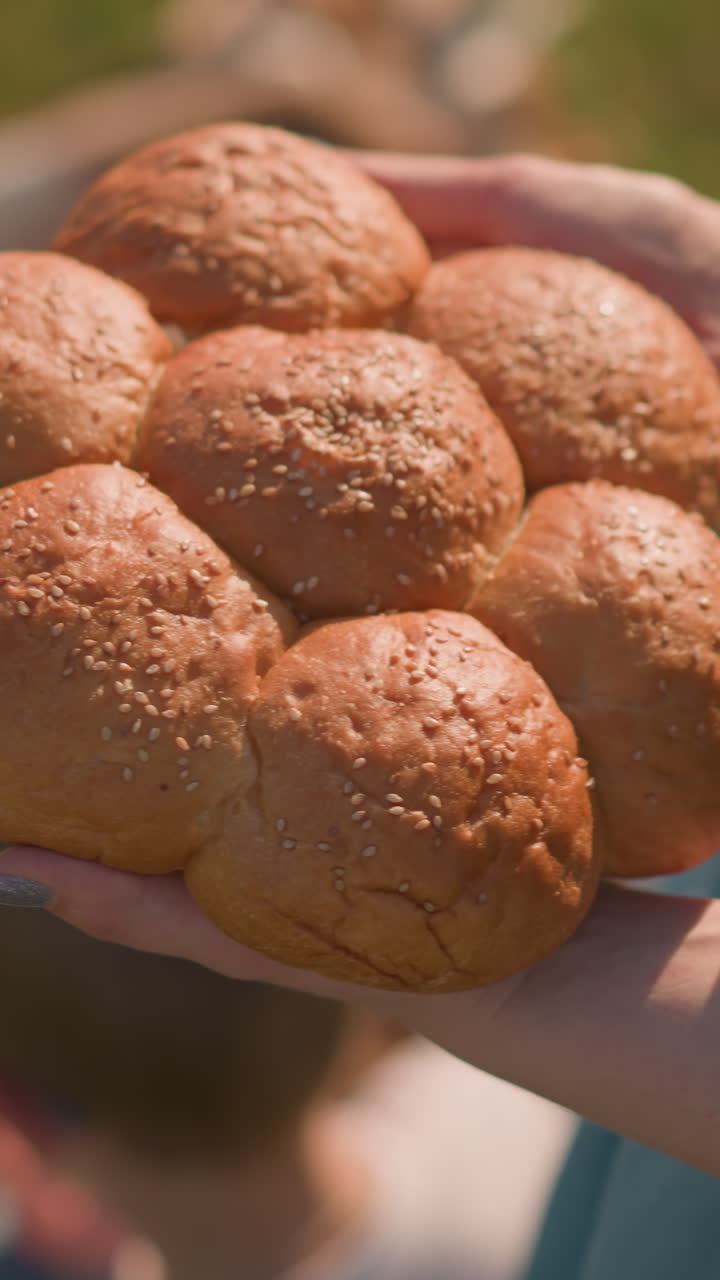 una mujer con un vestido azul sostiene un grupo de rollos de pan de semillas de sésamo frescos, con sus hijos borrosos en el fondo. el primer plano se centra en el pan