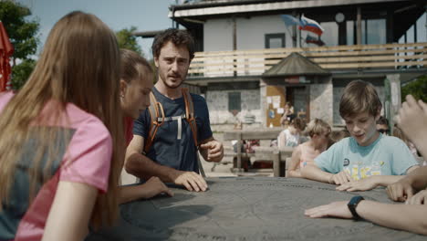 hiker fastens his backpack and chating with the kinds who are gathered around a round table with a map of directions for the world cities and destinations