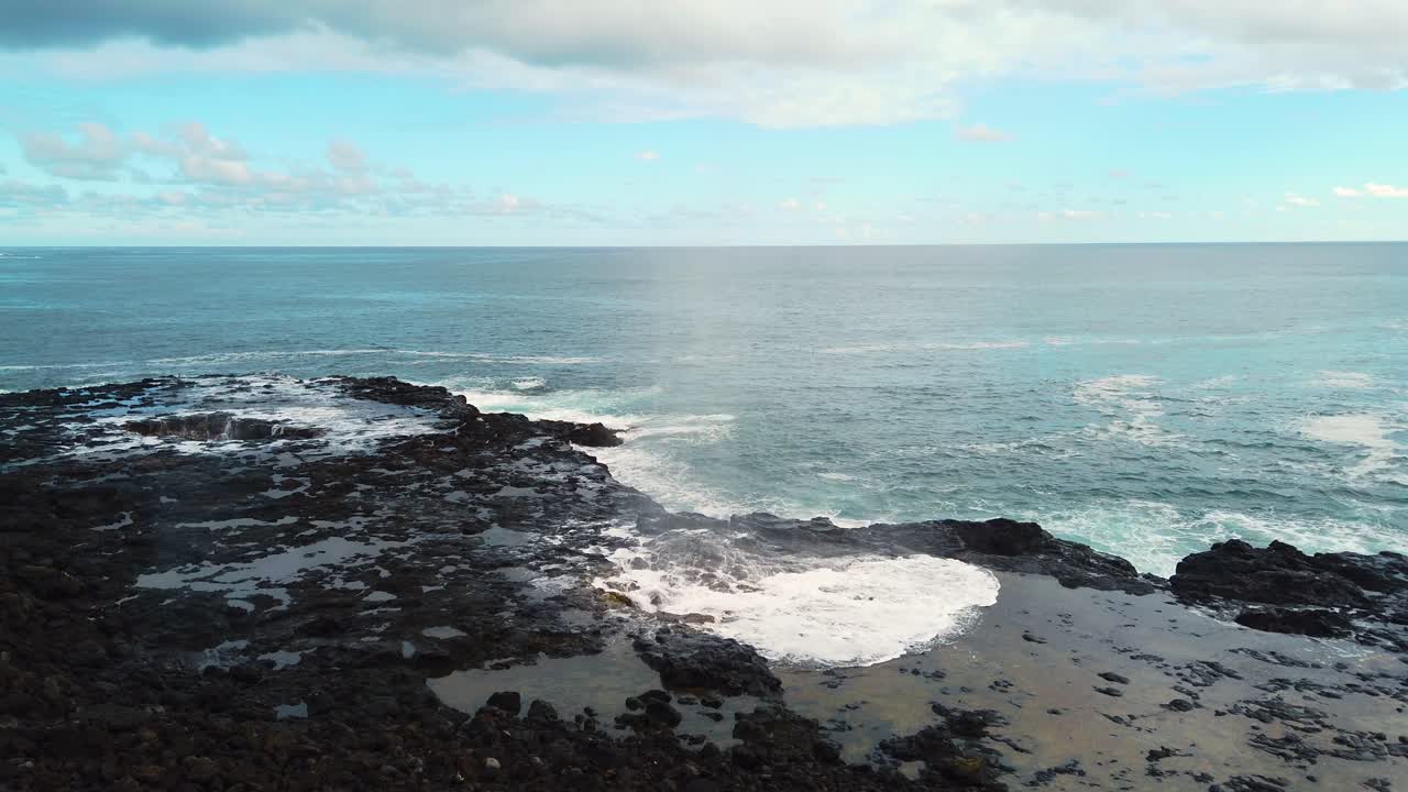 4K Hawaii Kauai static Spouting Horn blowhole going off twice in center of frame with water in hole in lava rock in left of frame with ocean waves in distance with partly cloudy sky