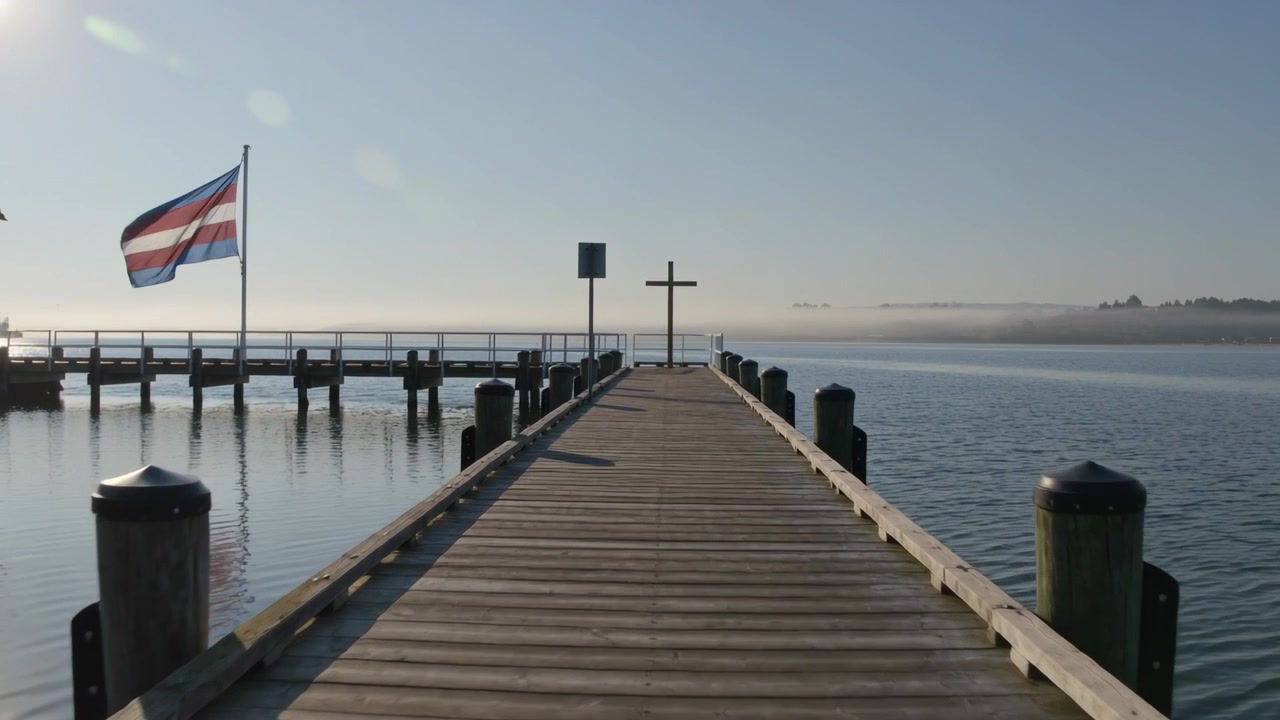 La telecamera inizia a riprendere, seguendo un molo di legno che si estende in una baia calma, per mettere in risalto un palo a croce con una bandiera che sventola.