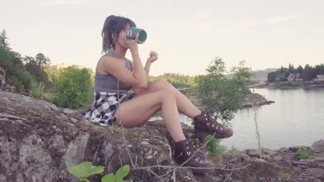 cute hipster girl drinking from a mug in front of the water in a park