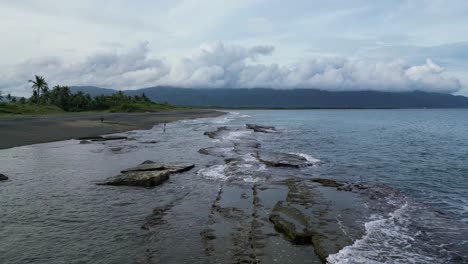 Toma-Aérea-De-Drones-De-Olas-Rompiendo-En-Rocas-Costeras-Mientras-La-Niña-Camina-En-La-Playa