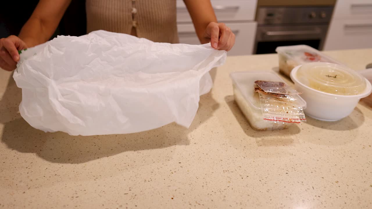 A woman in a kitchen handles a plastic bag over a counter with food containers, emphasizing recycling and sustainability