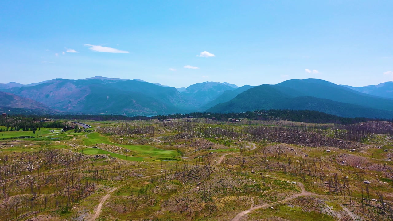Drone panorama of a golf course carved into wildfire aftermath, with bright fairways, scorched hillsides, scattered dead timber, curving cart paths, and layered mountains on the horizon.