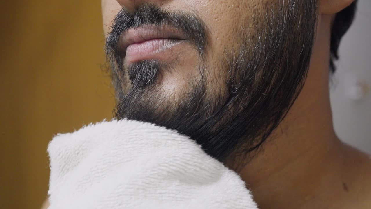 A closeup footage of a young man drying his beard with a white towel, with blurred background. Hygiene concept