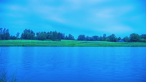 Cinematic-night-to-day-timelapse-of-the-sun-rising-over-a-beautiful-and-calm-lake-with-trees-and-green-grass-in-the-background-on-a-cloudy-day