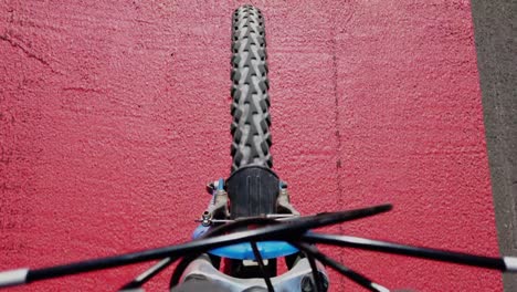 point of view pointing down on a bikeride, showing the front tire, the ground is the bright red marking of a bycicle path in a city