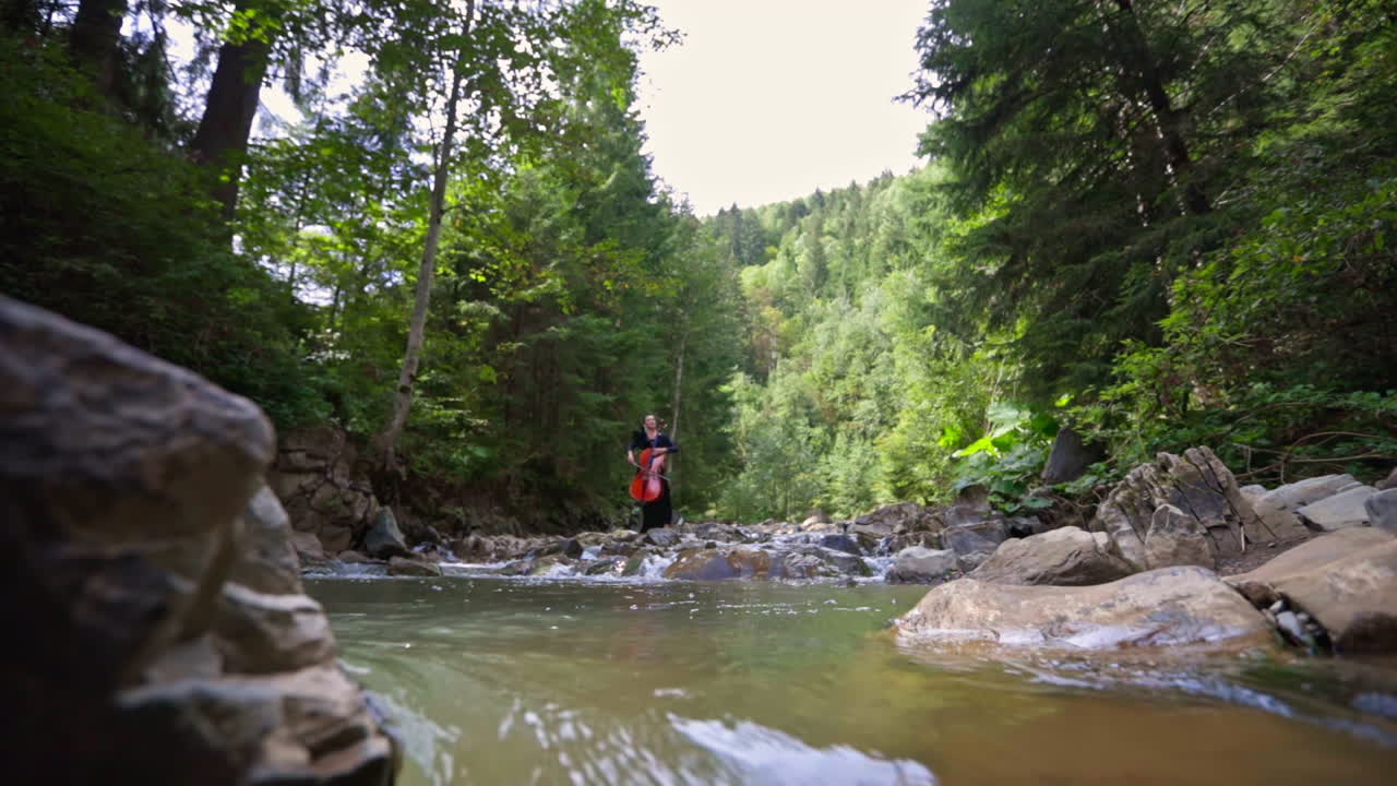 Female cellist at the river. Woman playing the cello on green trees background. Fresh mountain water flowing among stones.