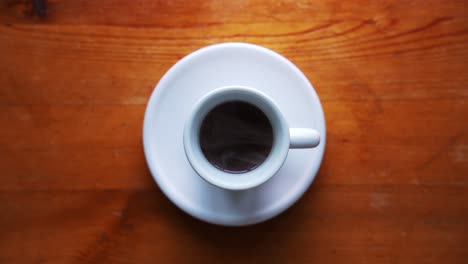 down shot o a hot espresso cup standing on a white plate on a brown wooden table