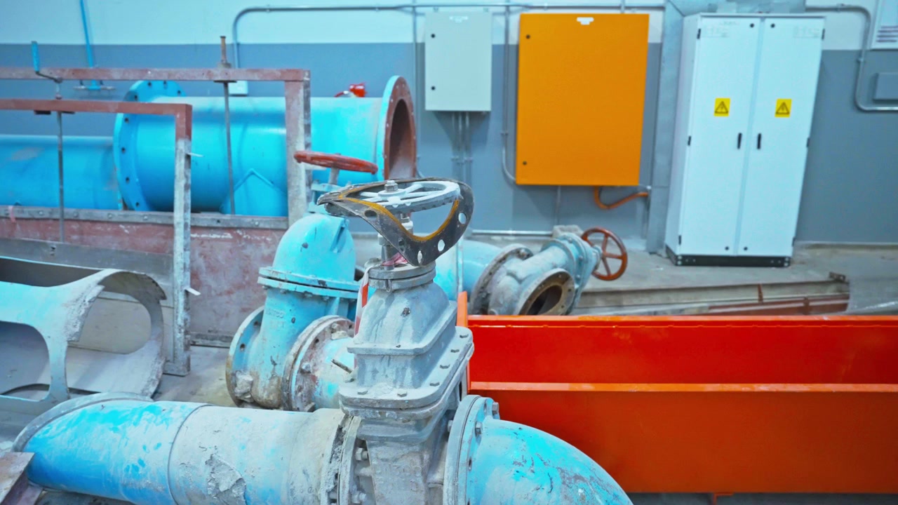 Interior view of an industrial water treatment or pumping facility showing large blue pipes, manual gate valves with red handwheels, electrical control panels, and an orange steel trough.