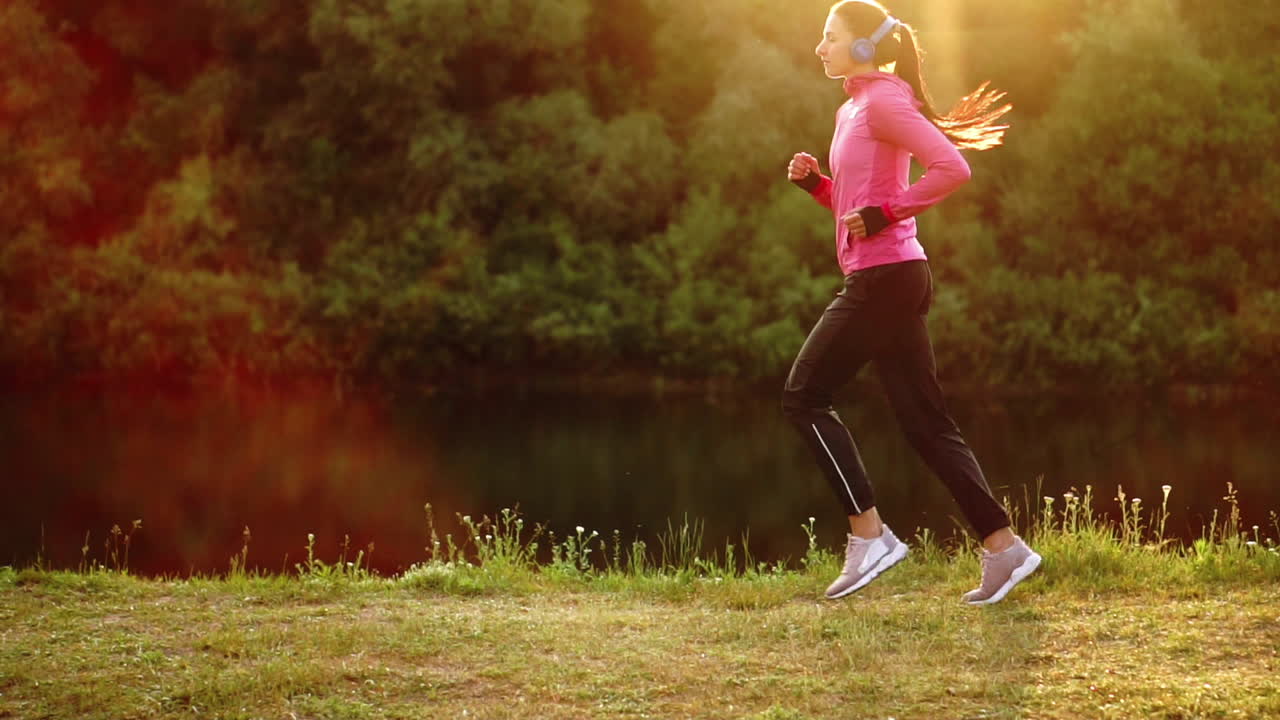 Brunette with long hair in headphones runs along the river in the Park in the morning at sunrise in the summer in a pink jacket and black pants