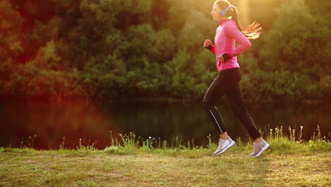 brunette with long hair in headphones runs along the river in the park in the morning at sunrise in the summer in a pink jacket and black pants