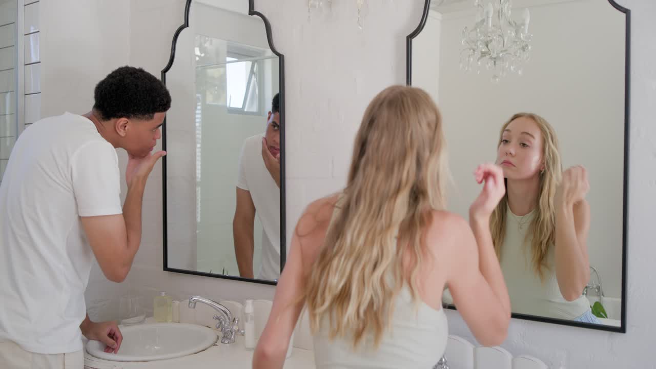 Coppia multietnica che si prepara per la giornata: l'uomo si sciacqua il viso mentre la donna applica la crema alla toeletta di casa.