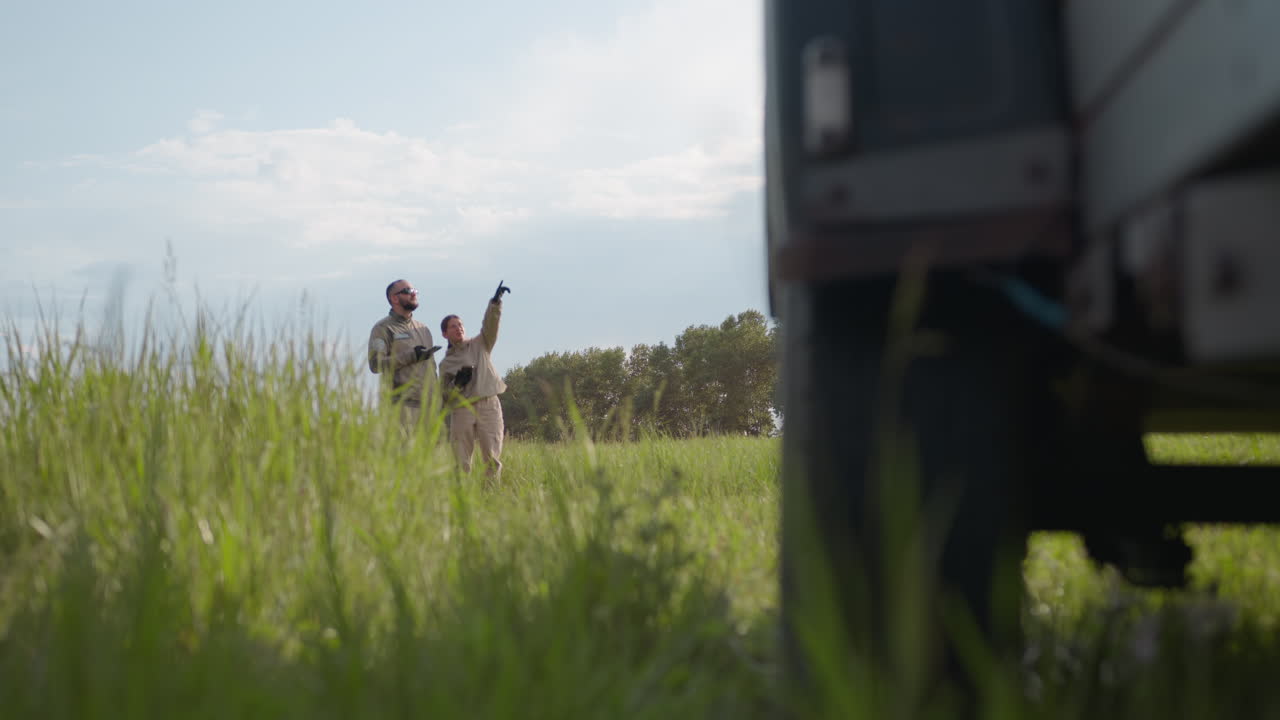 close up of parked car side and wheel in foreground with woman pointing skywards at distant object while male partner watches attentively amid tall grass and trees under cloudy blue sky