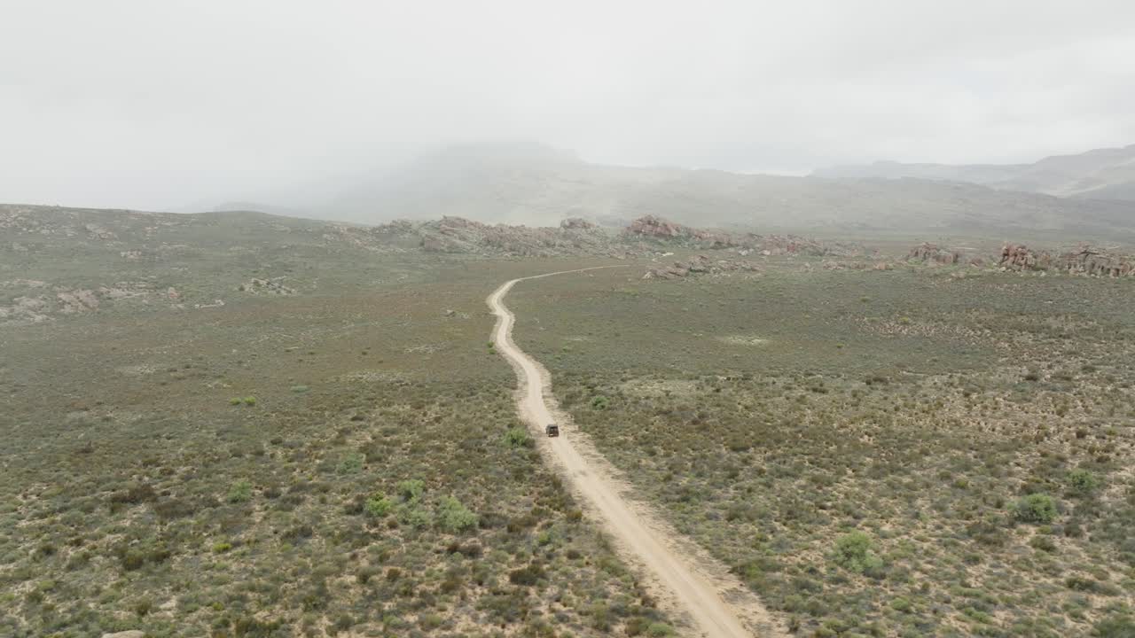 drone vuela alto sobre el paisaje desértico sobre una carretera donde un vehículo fuera de carretera negro conduce en el área desértica de cederberg en sudáfrica - en el fondo se pueden ver montañas rocosas