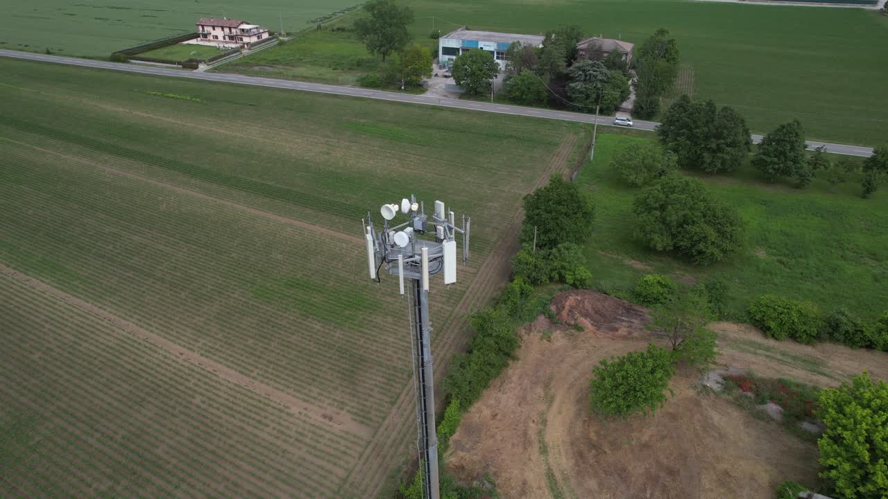 Rural cellular base station tower stands beside farmland with sector panel antennas, microwave backhaul dishes, and a fenced equipment area near country roads and trees under soft light, drone orbit